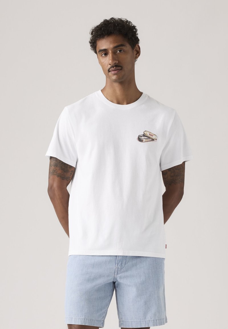 Man met tatoeages, draagt een wit t-shirt met een klein afbeelding op de borst en lichtblauwe spijkershorts, staand tegen een effen achtergrond.