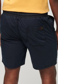 Hombre con camiseta amarilla y pantalones cortos azul marino con cinturilla elástica y bolsillo trasero con una pequeña etiqueta en el lado derecho.
