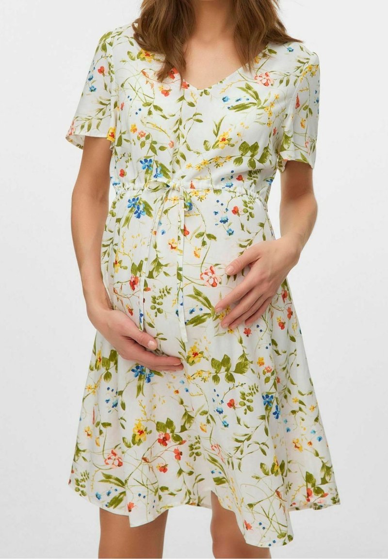 Femme enceinte portant une robe blanche à fleurs avec des manches courtes, caressant doucement son ventre avec ses deux mains.