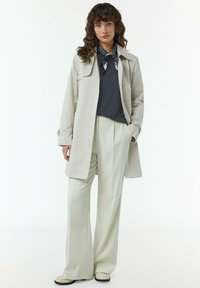 Jasno beżowy trencz na granatowej bluzce, zestawiony z spodniami w kolorze kremowym wysokim w staniku oraz jasnymi butami. Wzorzysty szalik dopełnia stylizację.