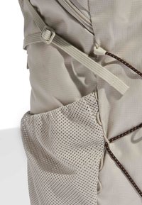 Zaino beige di lato che mostra la fascia regolabile, una tasca in rete e un dettaglio in corda elastica nera-arancione su tessuto texturizzato.