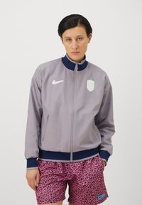 Svijetlosiva bomber jakna s mornarskim ovratnikom, koja ima bijeli Nike logo i grb, uparena s kratkim hlačama s ružičastim printom leopard uzorka.