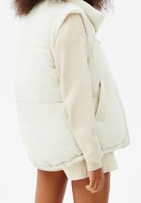 Gilet en crème duveteux avec col montant, poches latérales et cordon de serrage à l'ourlet, porté sur un sweatshirt beige clair. Texture douce.