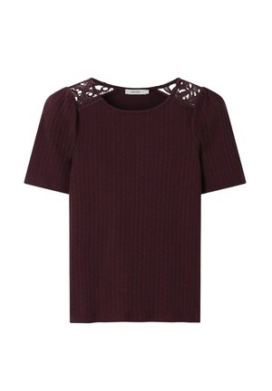 Femme portant de grandes lunettes de soleil marron, un haut bordeaux à manches courtes avec des détails en dentelle aux épaules, un pantalon rose clair et une ceinture marron.