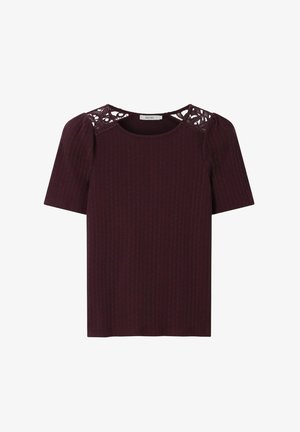 Femme portant de grandes lunettes de soleil marron, un haut bordeaux à manches courtes avec des détails en dentelle aux épaules, un pantalon rose clair et une ceinture marron.