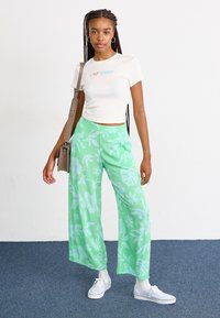 T-shirt blanc court avec du texte bleu et rouge, associé à un pantalon large vert avec un motif floral blanc, et des baskets blanches.