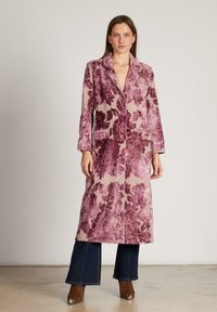 Abrigo largo rosa con patrón floral oscuro, hecho de un tejido texturizado, que presenta un collar, botones frontales y dos bolsillos en el pecho. Combinado con jeans de pierna ancha.