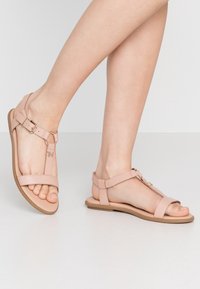 Vaaleanpunaiset litteät sandaalit, joissa on remmit, säädettävä solki ja T-muotoinen muotoilu. Pehmeä materiaali ja kumipohja mukavuutta ja joustavuutta varten.