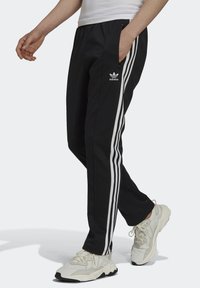 Pantaloni tuta neri con accenti bianchi a tre strisce e logo Adidas, caratterizzati da una vestibilità tapered e tasche laterali con zip. Indossati con sneakers chiare.