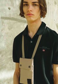 Svart polo skjorte med knappet krage, som har en beige lomme med trykknapper og skulderrem. Enkelt tekstur, minimalistisk design.