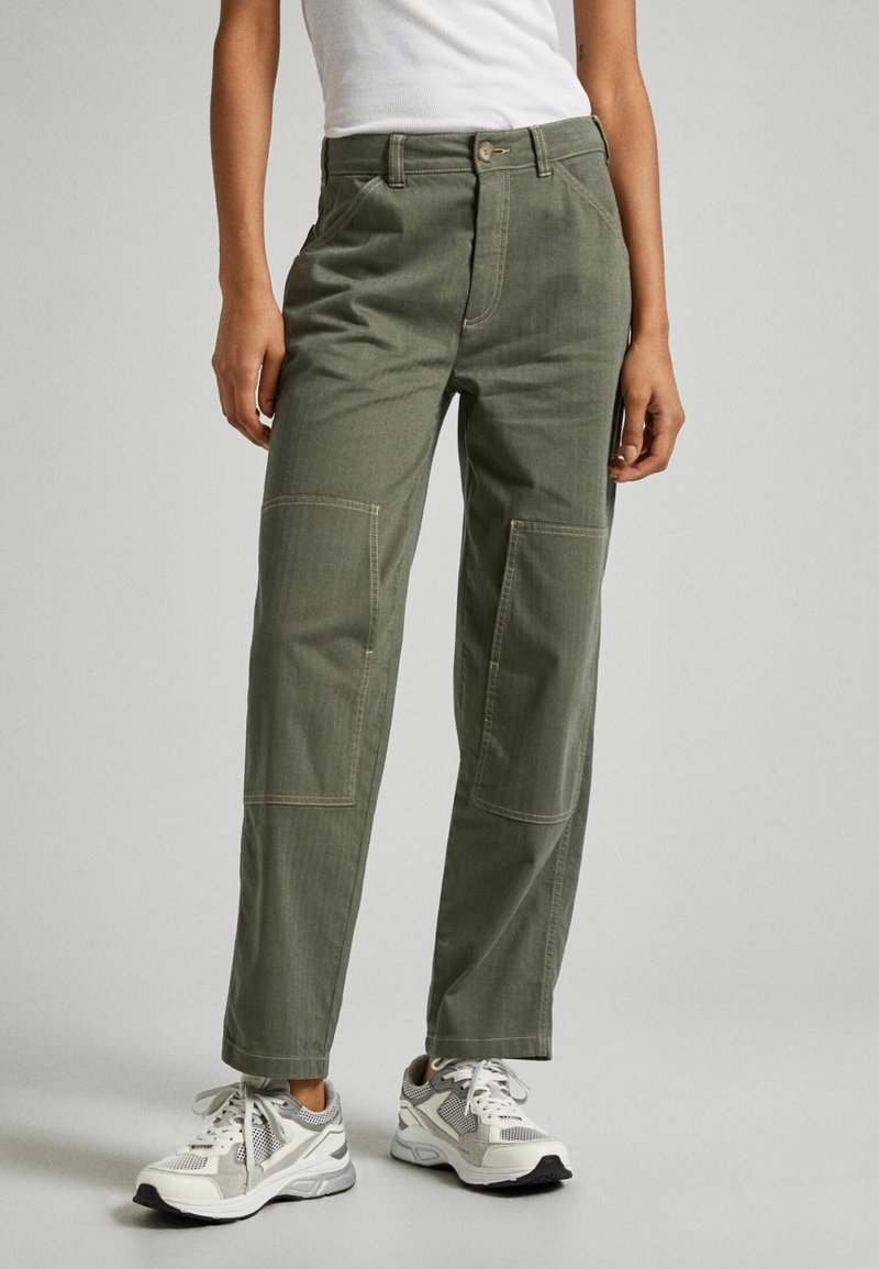 Personne portant un pantalon droit kaki avec des poches plaquées à l'avant, un haut blanc sans manches, et des chaussures de sport blanches et grises.