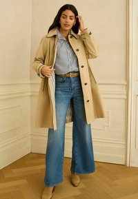 Femeie purtând o trench bej peste o cămașă în dungi, blugi evazați albaștri și cizme elegante bej cu toc, stând pe un podea de lemn în fața unui perete bej.