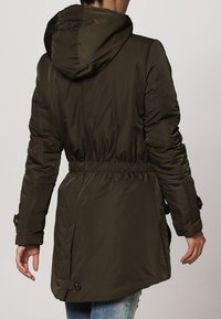 Veste matelassée vert olive avec une taille élastique, une capuche et un dos légèrement plus long. Présente des coutures visibles et des accents de boutons.