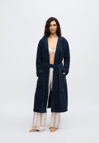 Βαθύ μπλε ρόμπα με υφή, με κουκούλα και ζώνη, με δύο μπροστινές τσέπες, φορεμένη πάνω από ανοιχτόχρωμο, ριγέ παντελόνι.