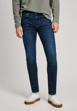 Jeans in denim blu scuro con vestibilità slim, cinque tasche e dettagli di cucitura visibili. Abbinati a sneakers bianche con accenti color cammello.