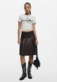 Béžové grafické tričko s púštnou scenériou, hnedá kožená plisovaná sukňa, čierne kovbojské čižmy so striebornou výšivkou a malá čierna kabelka.