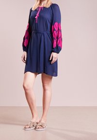 Mörkblå klänning med rosa broderade blommönster på ärmarna, insnörad midja och knytning i halsen. Modellen bär guldfärgade sandaler.