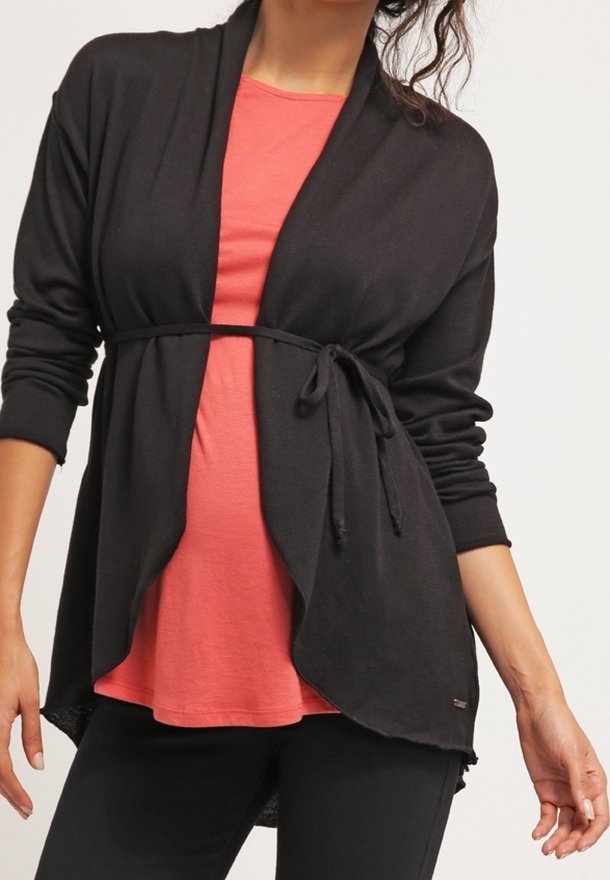 Femme portant une chemise corail sous un cardigan noir ouvert devant, noué à la taille, associé à un pantalon noir, sur un fond uni.
