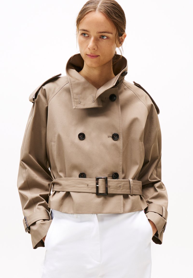 Manteau en beige, coupé et léger, doté de boutons double boutonnage, d'une taille ceinturée et d'un col montant avec épaulettes.