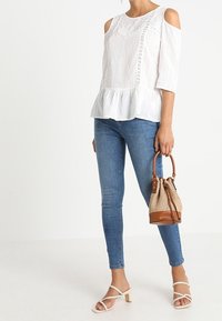 Blusa bianca a spalle scoperte con tessuto testurizzato e dettagli in pizzo, orlo peplum, abbinata a jeans skinny in denim blu e una borsa a secchiello marrone.