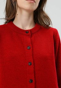 Cardigan rouge en maille avec un col rond, comportant une rangée de cinq boutons noirs sur le devant. Texture douce et coupe confortable.