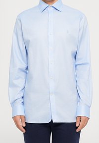 Man som bär en ljusblå långärmad skjorta med knappar och krage, i kombination med mörkblå byxor, stående mot en neutral bakgrund.