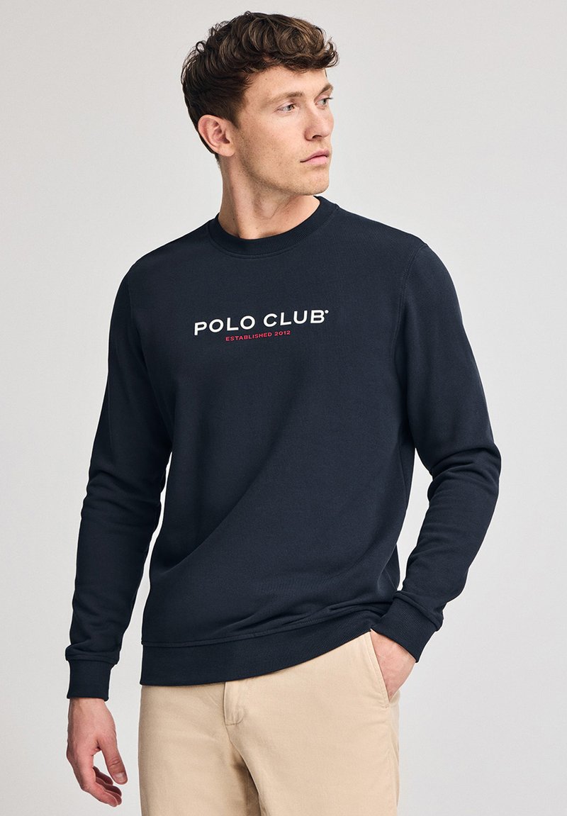 Marinblå sweatshirt i ett slätt tyg, med "POLO CLUB"-logotyp och "ETABLERAD 2012" i vit och röd text. Klassisk rundhalsad design.