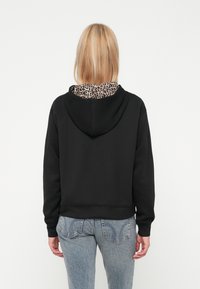 Svart kortermet hettegenser med leopardmønster på innsiden av hetten. Har en myk tekstur og lange ermer, kombinert med lyseblå jeans.