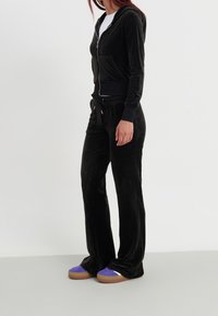 Chándal de terciopelo negro con sudadera con cierre y pantalones con cordón. Presenta una textura suave y un ajuste relajado, combinado con zapatillas moradas y beige.