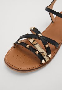 Sandale en cuir noir avec accents de clous dorés, présentant une bande à imprimé léopard texturé. Semelle plate avec perforations pour la respirabilité.