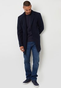 Marineblauer Wollmantel mit Reverskragen, getragen über einem dunklen Strickpullover, kombiniert mit blauen Jeans und dunklen Sneakers. Einfaches Design, ohne Muster.