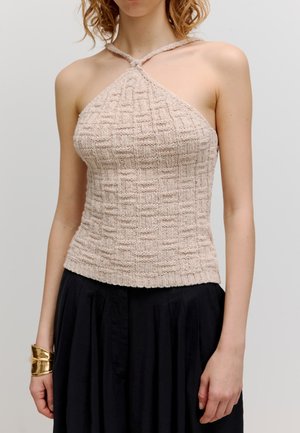 Strick-Haltertop in hellem Beige mit einem strukturierten Rautenmuster, ausgestattet mit einem geknoteten Träger und einer taillierten Silhouette. Kombiniert mit einem schwarzen Rock.