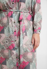 Robe à manches longues, boutonnée, présentant un design en patchwork avec des couleurs rose, gris et teal. Comprend une ceinture assortie et des boutons ronds verts.