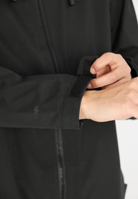 Zwarte zip-up jack met een gladde, matte afwerking; voorzien van een klittenbandsluiting op de manchetten en subtiele branding op de mouw.