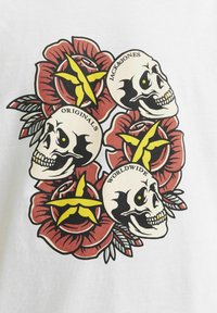 Hvid t-shirt med et grafisk design af fire kranier omgivet af røde roser og grønne blade, med gule stjerner og tekst accenter.