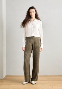 Witte geribbelde blouse met knopen, gecombineerd met losse olijfgroene broek met een subtiele luipaardprint. Het model staat op een houten vloer.