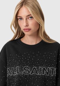 Czarna bluza z okrągłym dekoltem, ozdobiona srebrnymi ćwiekami tworzącymi napis "ALLSAINTS" w wyraźnej czcionce.