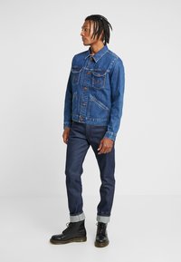 Niebieska kurtka dżinsowa o klasycznym designie, z dwoma kieszeniami na piersi, zapięciem na guziki i kontrastowym przeszyciem. W połączeniu z ciemnymi dżinsami i czarnymi bucikami.