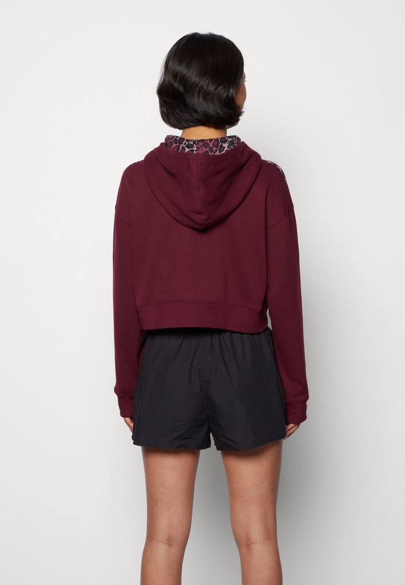 Kurzer burgunderfarbener Hoodie mit Kapuze und gemustertem Innenfutter, kombiniert mit schwarzen Shorts. Der Stoff wirkt weich, mit lockerem Schnitt und Ärmeln.