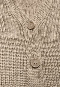 Vaaleanbeige neulecardigan, jossa on V-pääntie ja kaksi suurta pyöreää nappia sekä tekstuuroitu ribbikuvio. Pehmeä ja lämmin kangas.