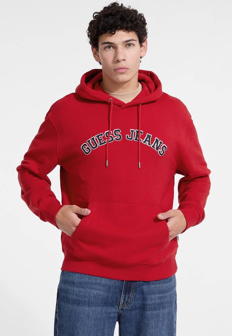 Červená mikina s kapucňou a predným vreckom, s šnúrkami na stiahnutie a vyšitou logom "GUESS JEANS" v bielej a čiernej farbe naprieč hrudníkom. Bavlnený materiál.