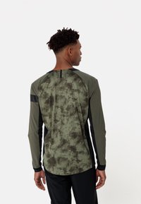 Chemise à manches longues de couleur vert foncé avec un motif tie-dye, panneaux latéraux noirs et col rond. Présente des coutures visibles et une fente au dos.