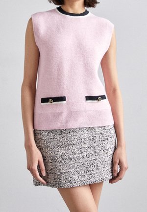 Mujer con top de punto sin mangas rosa con bolsillos abotonados en negro y blanco y una falda con patrón en blanco y negro, delante de un fondo liso.