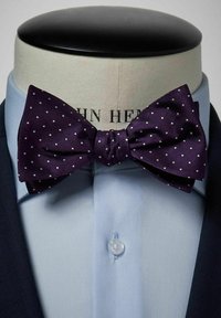 Lila prickig fluga av texturerad tyg, med en central knut, uppvisad på en mannekäng som bär en ljusblå skjorta och en mörk jacka.