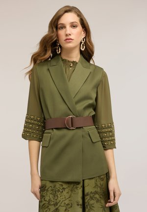 Donna con capelli lunghi e ondulati che indossa un vestito verde a strati con maniche decorate, ampia cintura marrone e grandi orecchini a cerchio dorati, sfondo neutro.