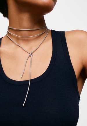 Persona que lleva una camiseta sin mangas negra y un collar plateado de varias capas con un diseño anudado alrededor del cuello y el pecho.