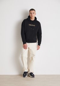 Felpa nera con tasca frontale e logo arancione, abbinata a pantaloni color crema e scarpe scure, su uno sfondo neutro.
