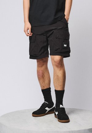 Mann trägt schwarze Cargo-Shorts, schwarze Socken und schwarze Turnschuhe, steht auf einer runden grauen Plattform vor einem schlichten Hintergrund.