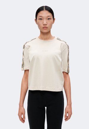 Jeune personne aux cheveux foncés attachés en arrière, portant un haut beige à manches courtes avec des rayures à motifs sur les épaules et un pantalon noir, face à l’avant.