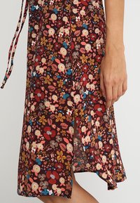 Abito midi floreale in un ricco tessuto bordeaux, caratterizzato da fiori e foglie colorati, con uno spacco laterale e sottili lacci in vita.